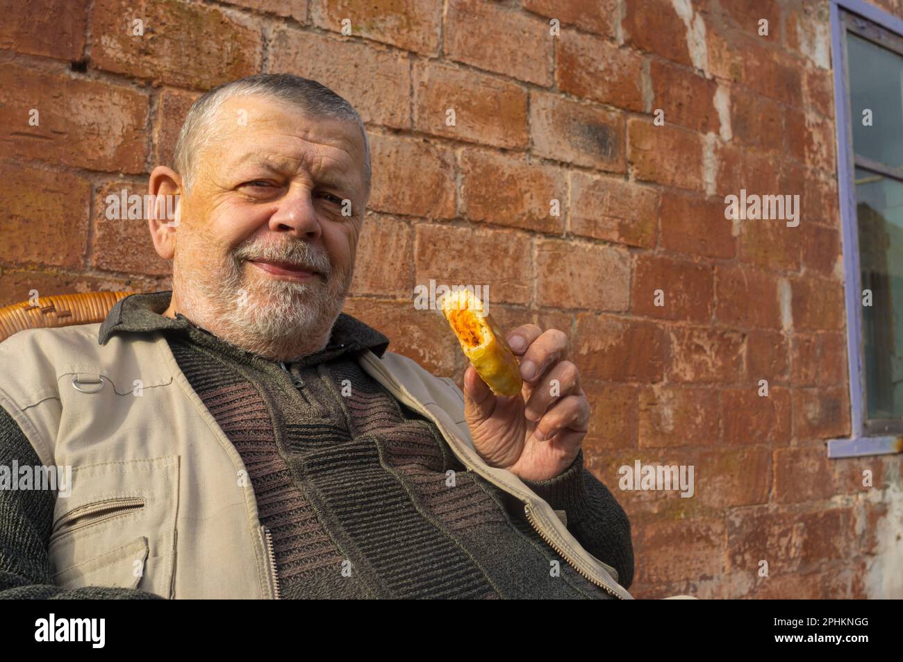 Ritratto all'aperto di un uomo anziano ucraino sorridente contro, mentre in piedi contro il mattone wal e tenendo in mano patty riempito di cavolo fritto Foto Stock