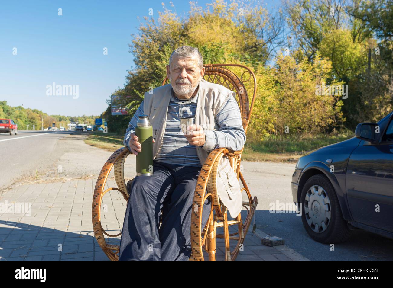 Ritratto dell'uomo anziano ucraino seduto in sedia a dondolo sul lato della strada e tenendo la bottiglia del vuoto che si prepara a bere il tè al giorno autunnale soleggiato Foto Stock