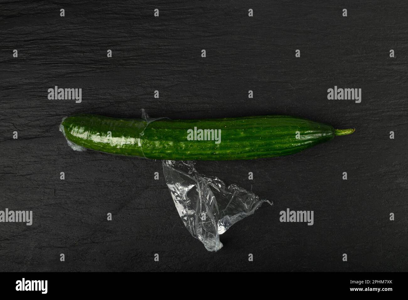 Cetriolo con involucro in plastica, imballaggio di verdure in pellicola, confezionamento di alimenti con involucro in pellicola, concetto di Eat Plastic, sfondo nero Foto Stock