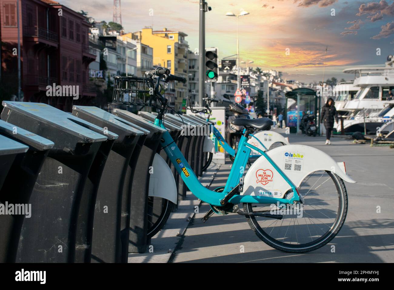 Servizio di noleggio biciclette, biciclette parcheggiate presso la stazione per biciclette. Un banner con le istruzioni su come noleggiare sullo sfondo e le persone che camminano sulla passerella. Foto Stock