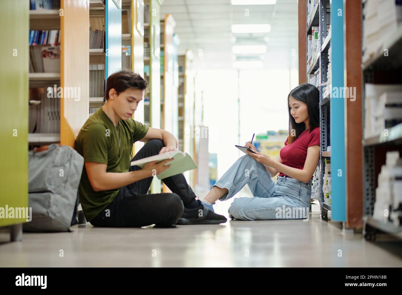 Studenti delle scuole superiori seduti al piano della biblioteca e leggendo un libro e lavorando al saggio Foto Stock