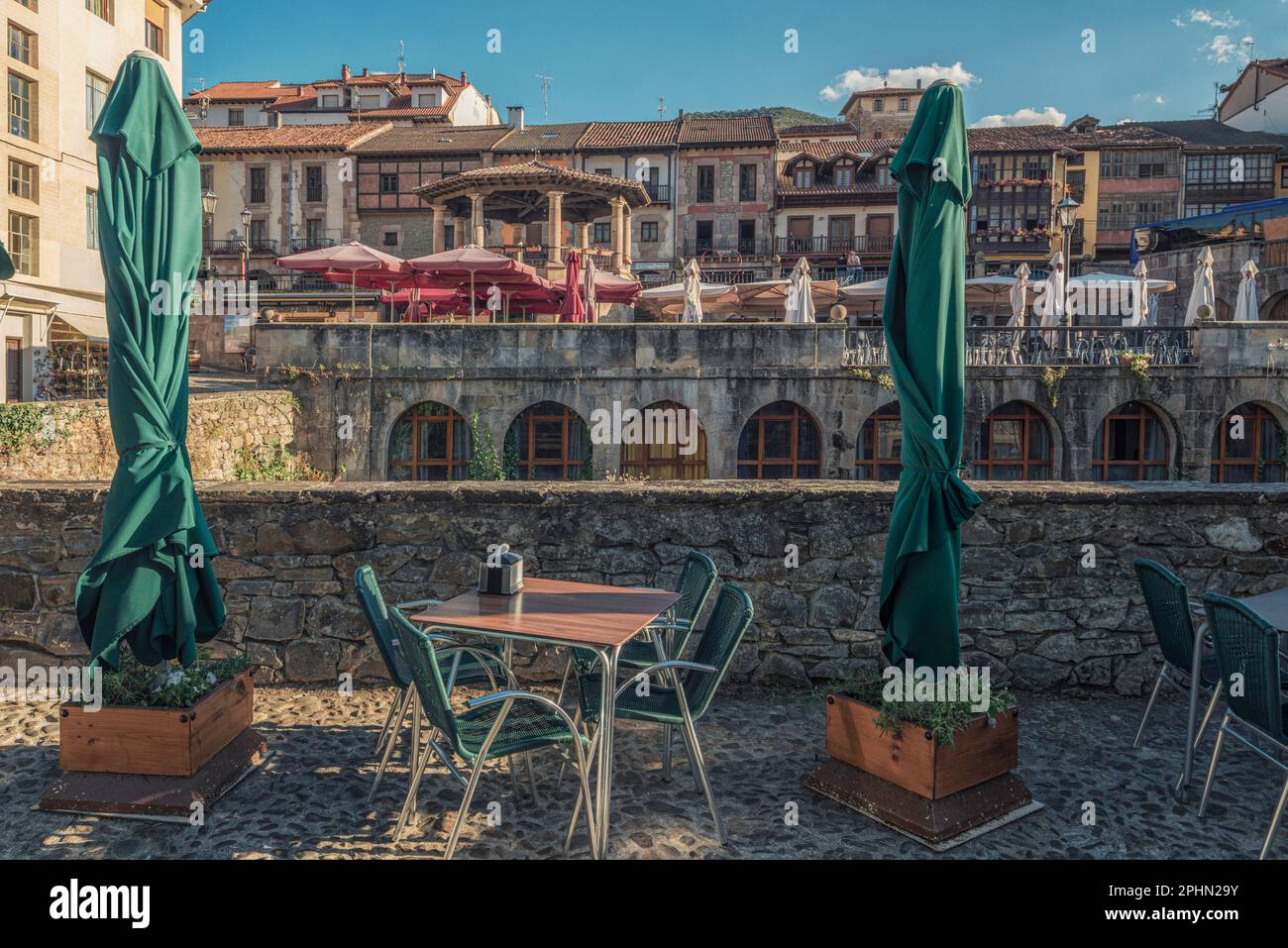 Terrazza con i suoi tavoli, sedie e ombrelloni sulla parete di pietra del fiume con la vista delle case e del tempio sullo sfondo, Potes, Foto Stock