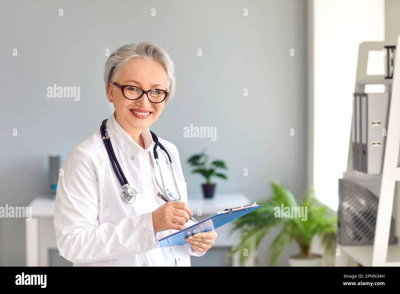 Felice medico senior con appunti in piedi in ufficio medico e sorridente Foto Stock