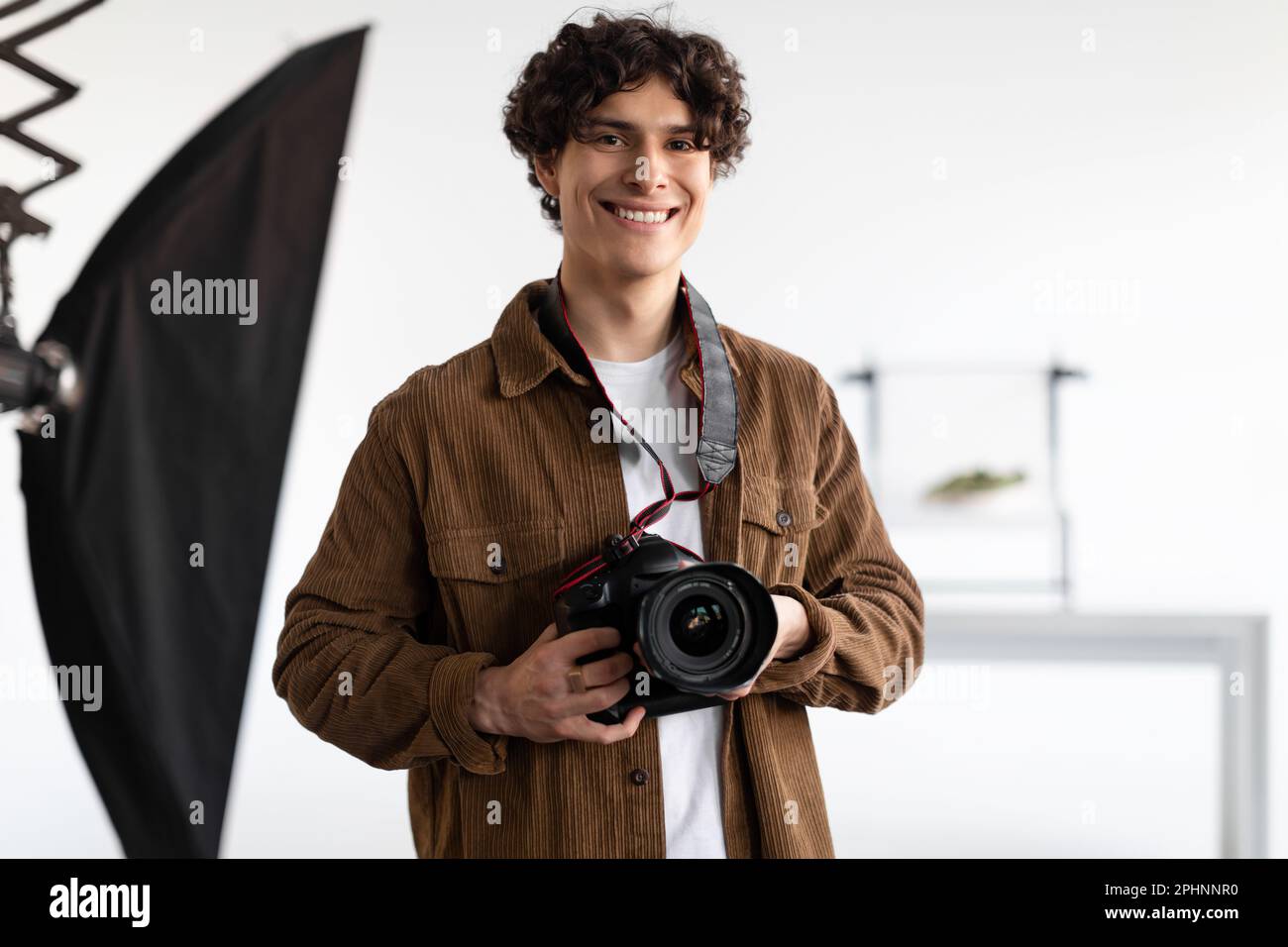 Felice giovane fotografo che tiene una macchina fotografica professionale, lavora in un moderno studio fotografico, sorridendo alla macchina fotografica Foto Stock