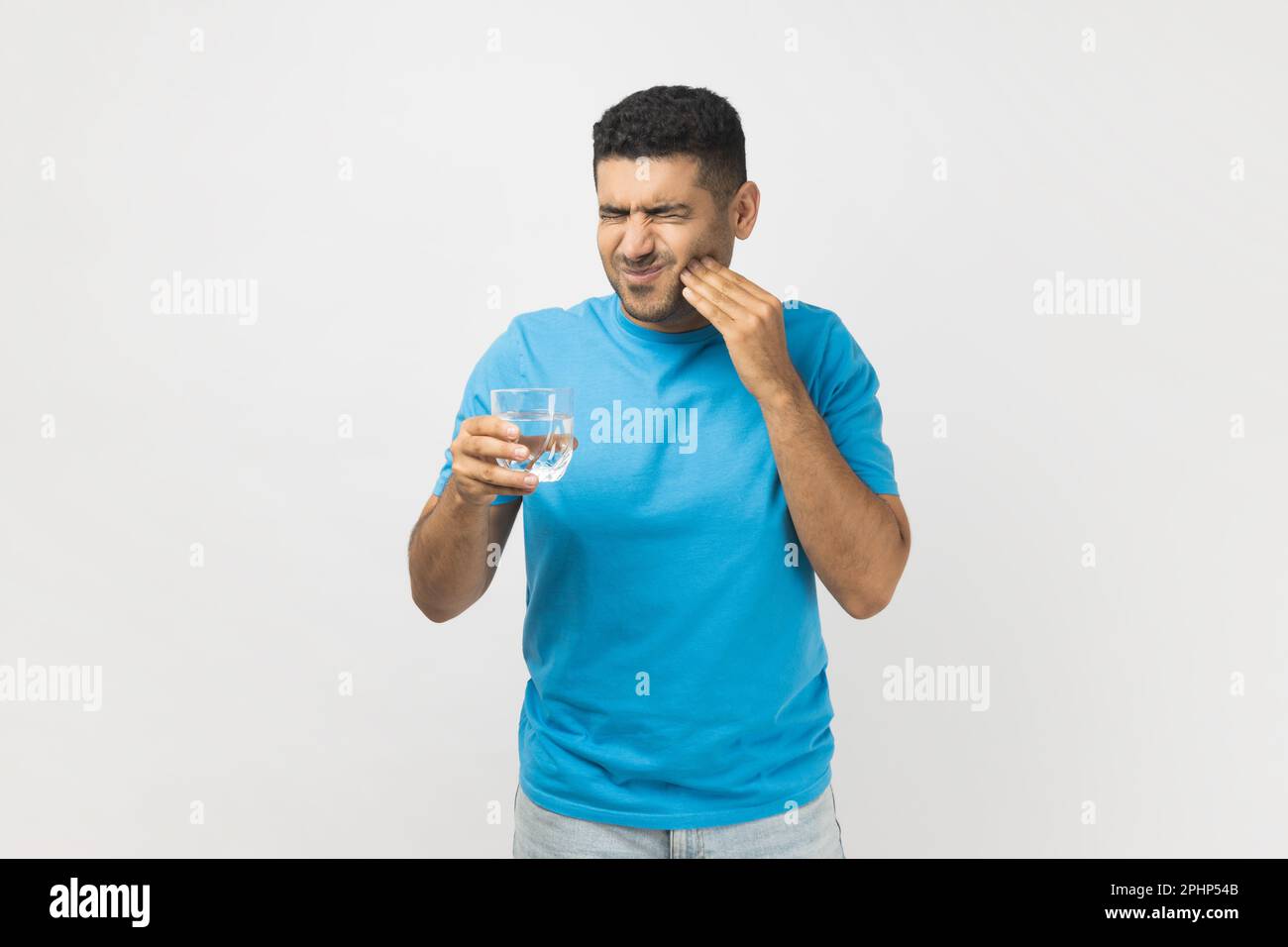 Ritratto di un uomo malsano non rasato che indossa una T-shirt blu in piedi con un bicchiere d'acqua fredda, sente dolore nel dente, ha denti sensibili. Studio in interni isolato su sfondo grigio. Foto Stock