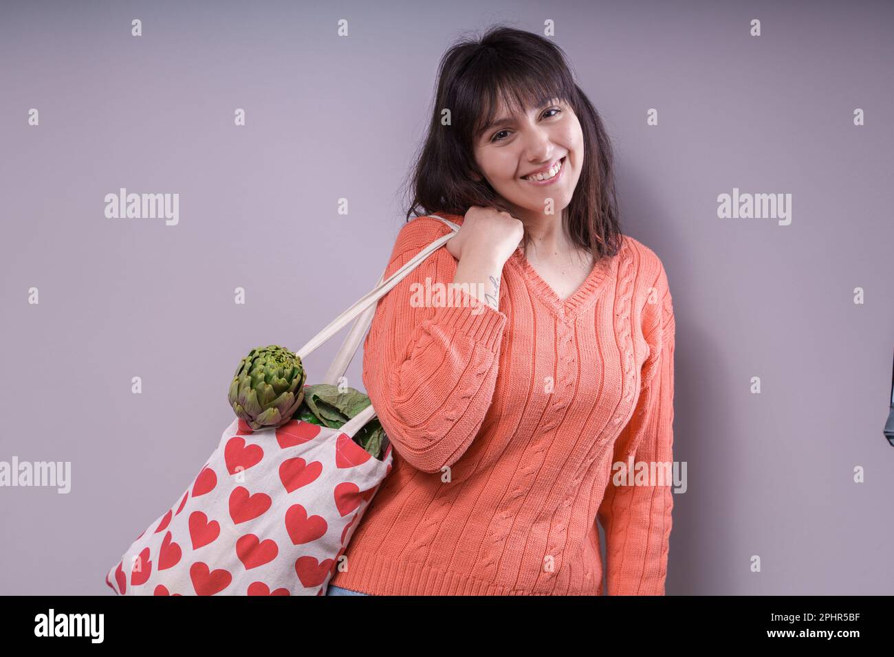 Giovane donna caucasica sorridente che tiene in mano una borsa per la spesa in tessuto con motivo cardiaco, ripiena di verdure (broccoli, cavolfiore, carciofo), guardando Foto Stock