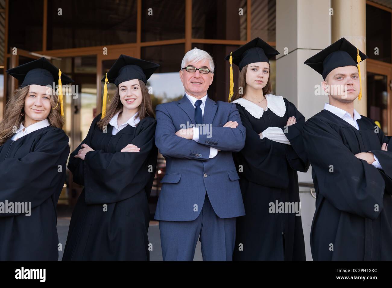 Un professore e i suoi studenti in abiti di laurea con le braccia incrociate sulle loro casse. Foto Stock