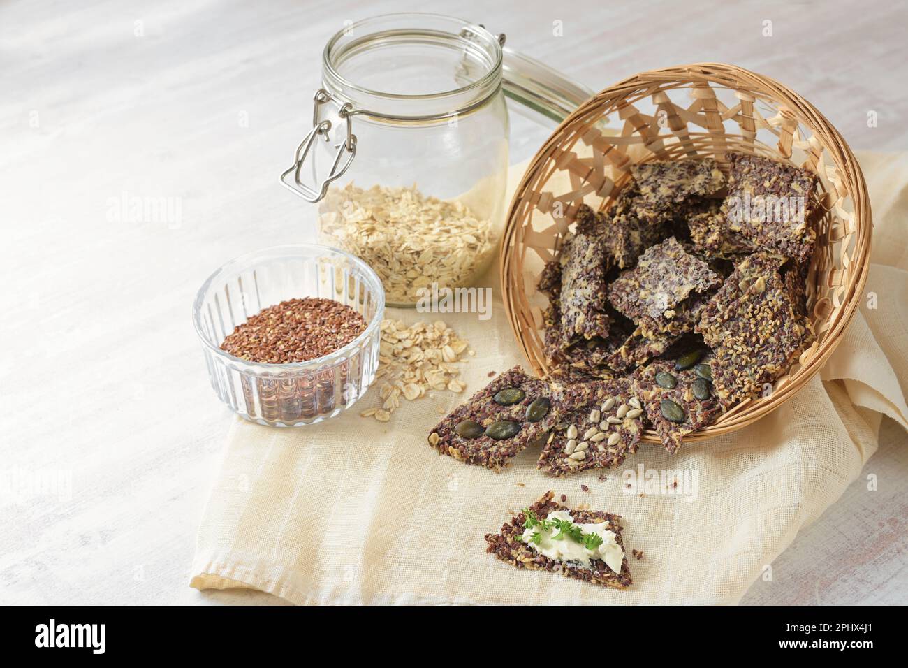 Snack croccanti di semi di lino, fiocchi di avena e vari semi in un cesto, uno con burro e prezzemolo guarnire, ingredienti in vasetti di vetro, tavolo leggero, co Foto Stock