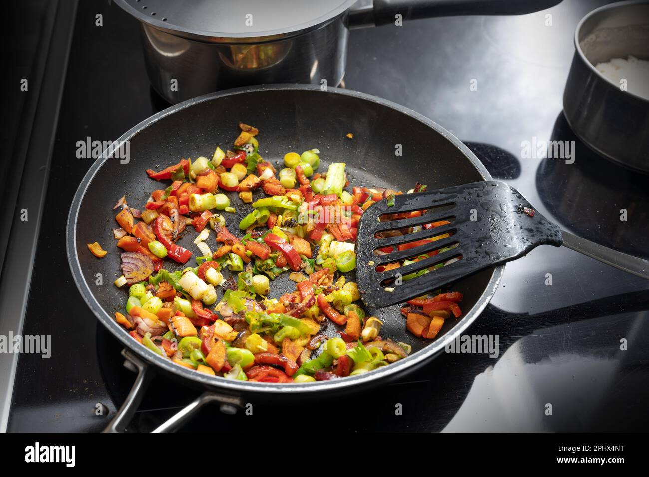 Mescolare le verdure fritte come il peperone, la cipolla e il porro con le spezie in una padella nera sul piano cottura con altre pentole, cucinando un curry vegetariano, Foto Stock