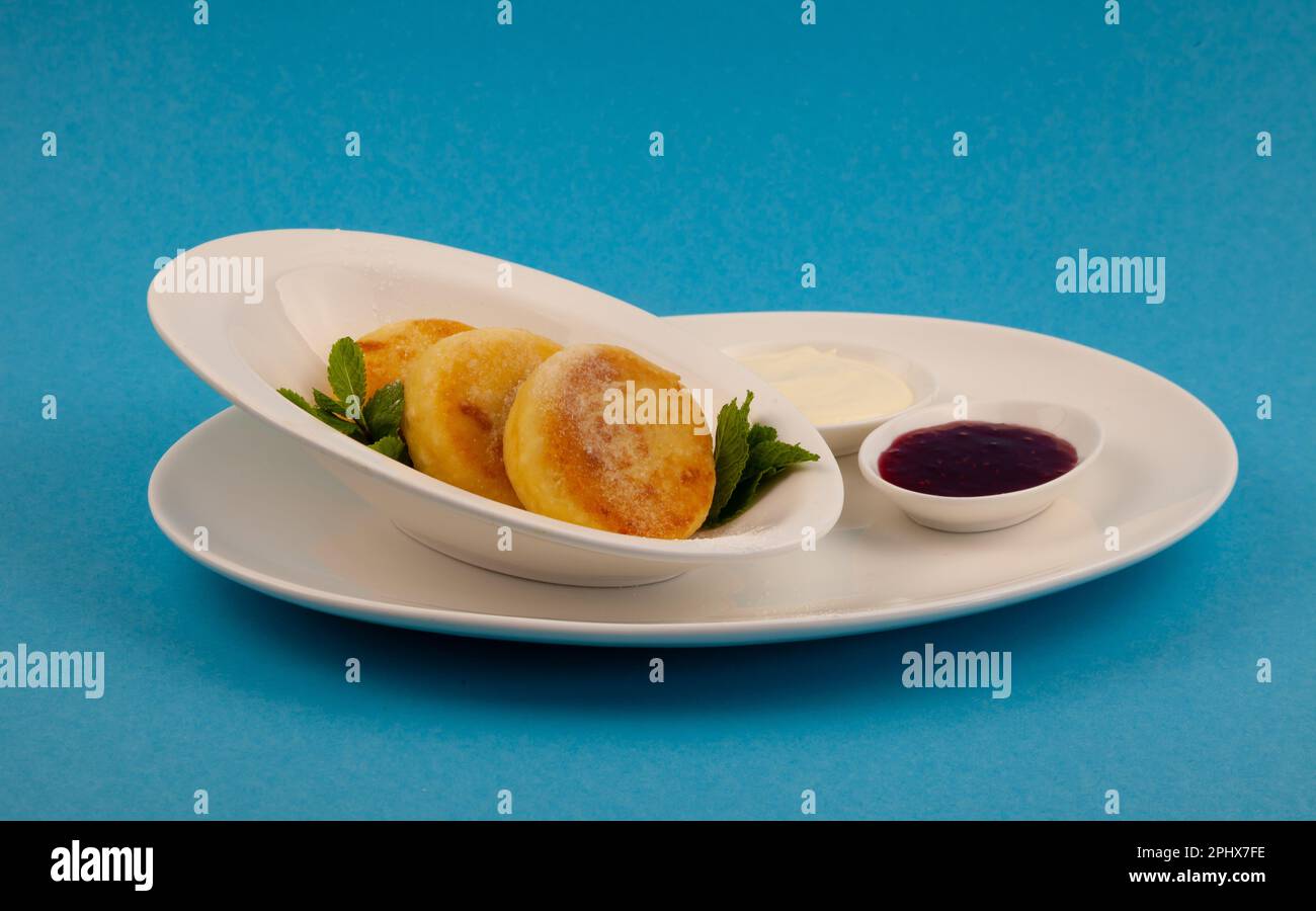 Frittelle di formaggio ricotta rotonda in un piatto bianco su sfondo blu. Cibo prima colazione. Cibo sano. Salsa di panna acida e menta. Primo piano vista dall'alto. Pasto dessert. Frittelle di ricotta al formaggio ricotta. Foto Stock