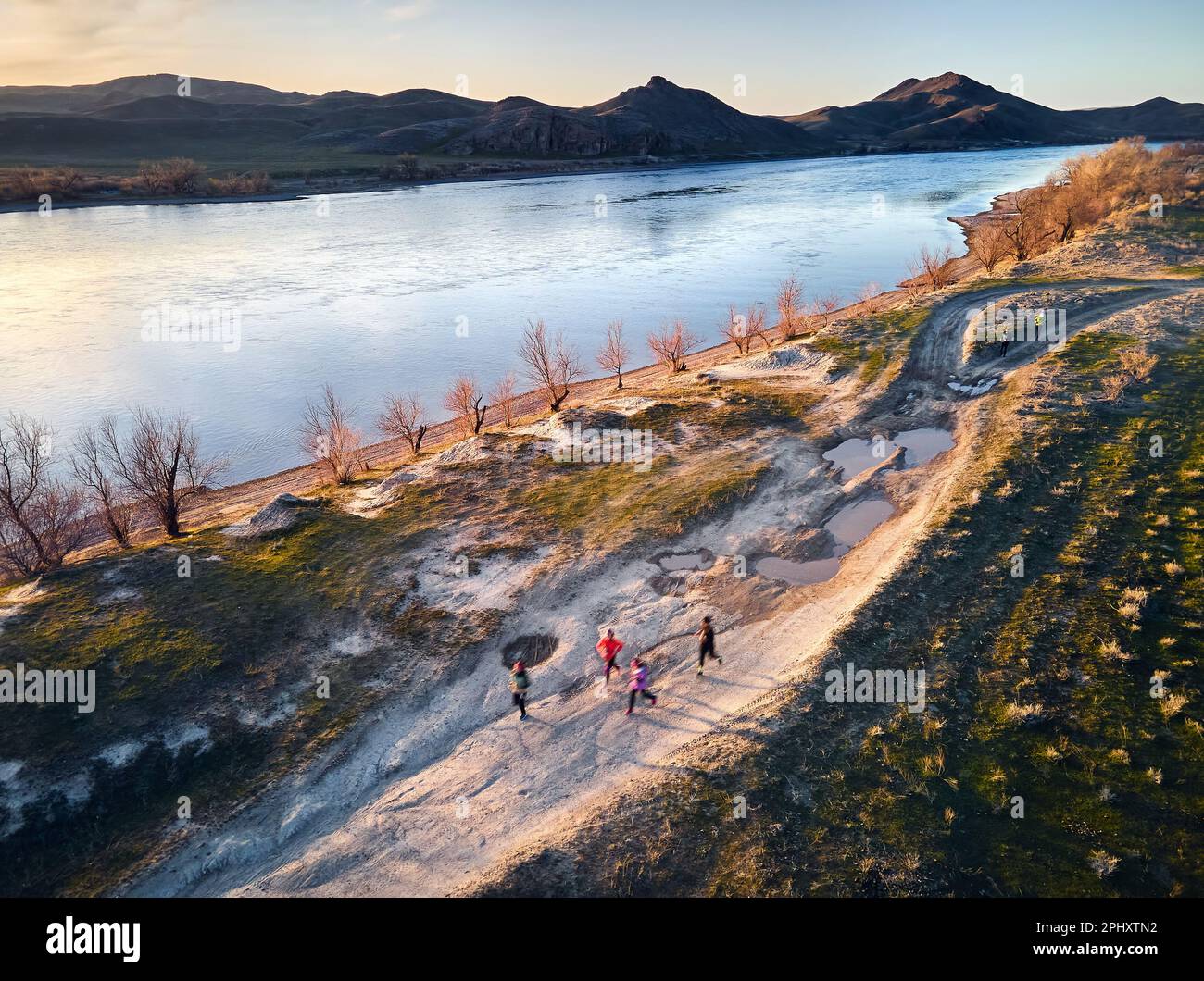 Vista aerea del drone dell'uomo atletico che corre lungo la strada di campagna polverosa vicino al fiume e alle colline al tramonto. Sentiero che corre alla steppa vicino al fiume Ili a sud Foto Stock