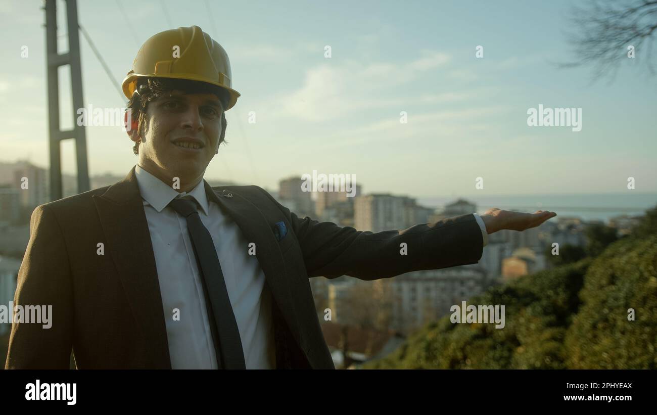 Professionista e sicuro giovane ingegnere o appaltatore in tuta e casco che mostra Condominiums moderni con una mano in Urban Landscap Foto Stock