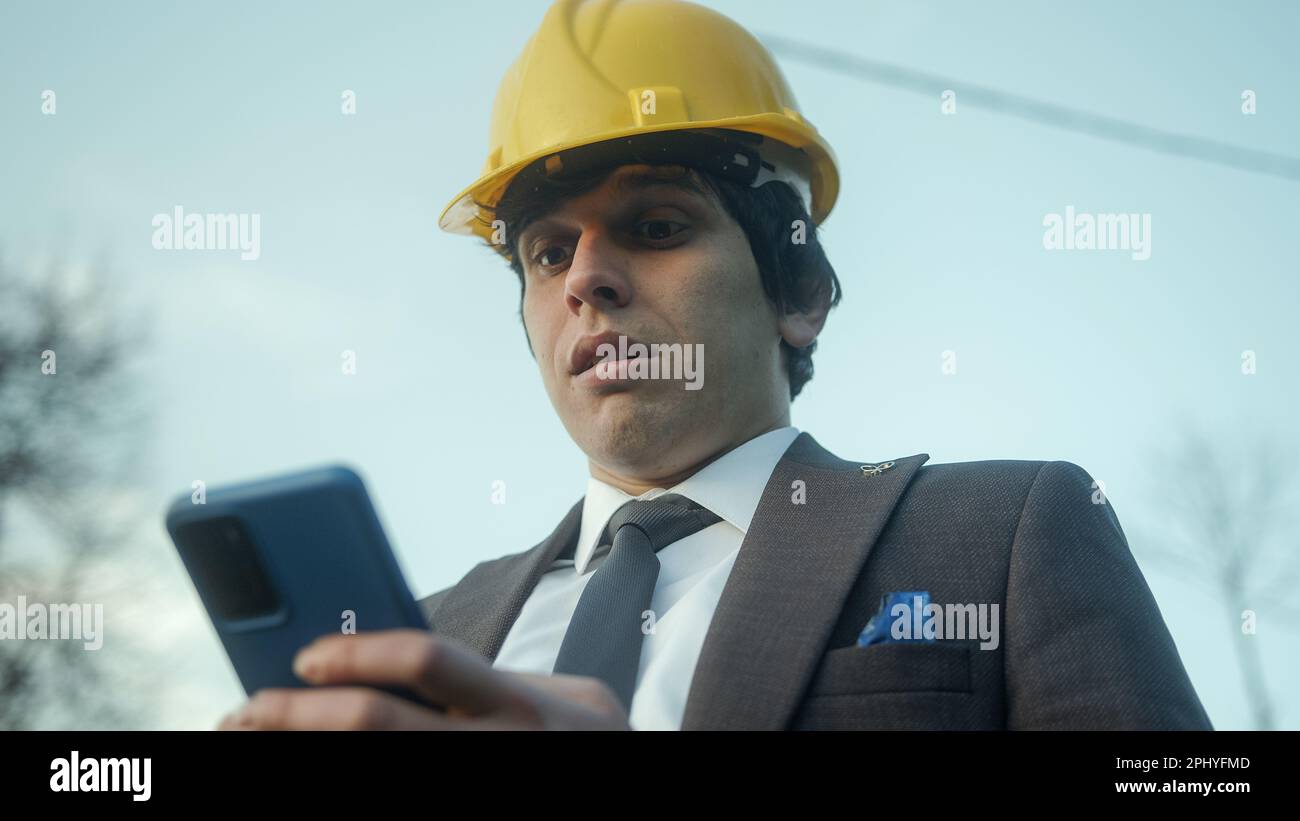 Sorpresa e gioia: Giovane ingegnere edile o uomo d'affari in casco utilizzando smartphone con gioia, viso sorpreso Foto Stock