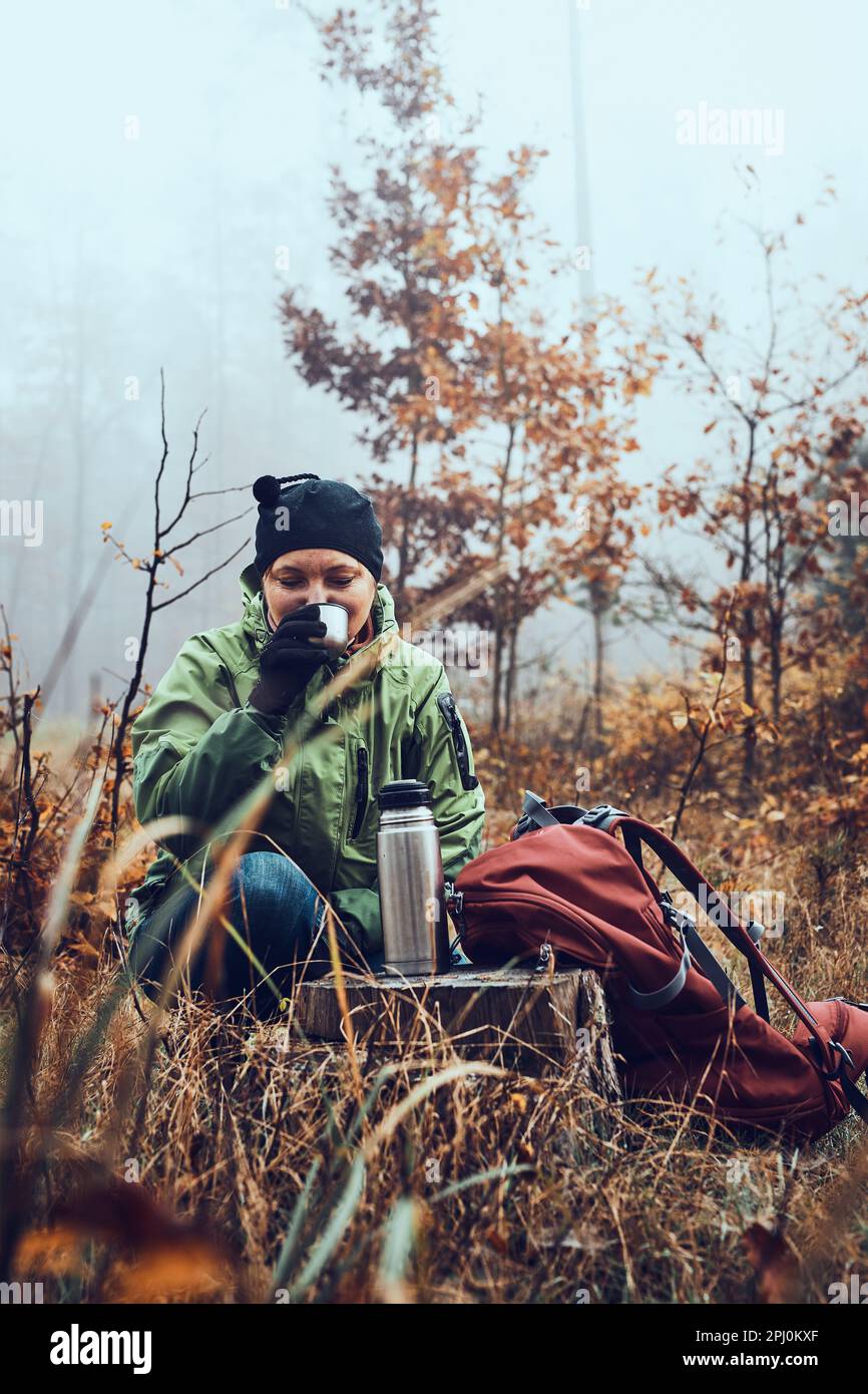 Donna con zaino in pausa durante il viaggio autunnale bere una bevanda calda da thermos beuta il giorno freddo autunnale. Attiva donna di mezza età che vagare arou Foto Stock