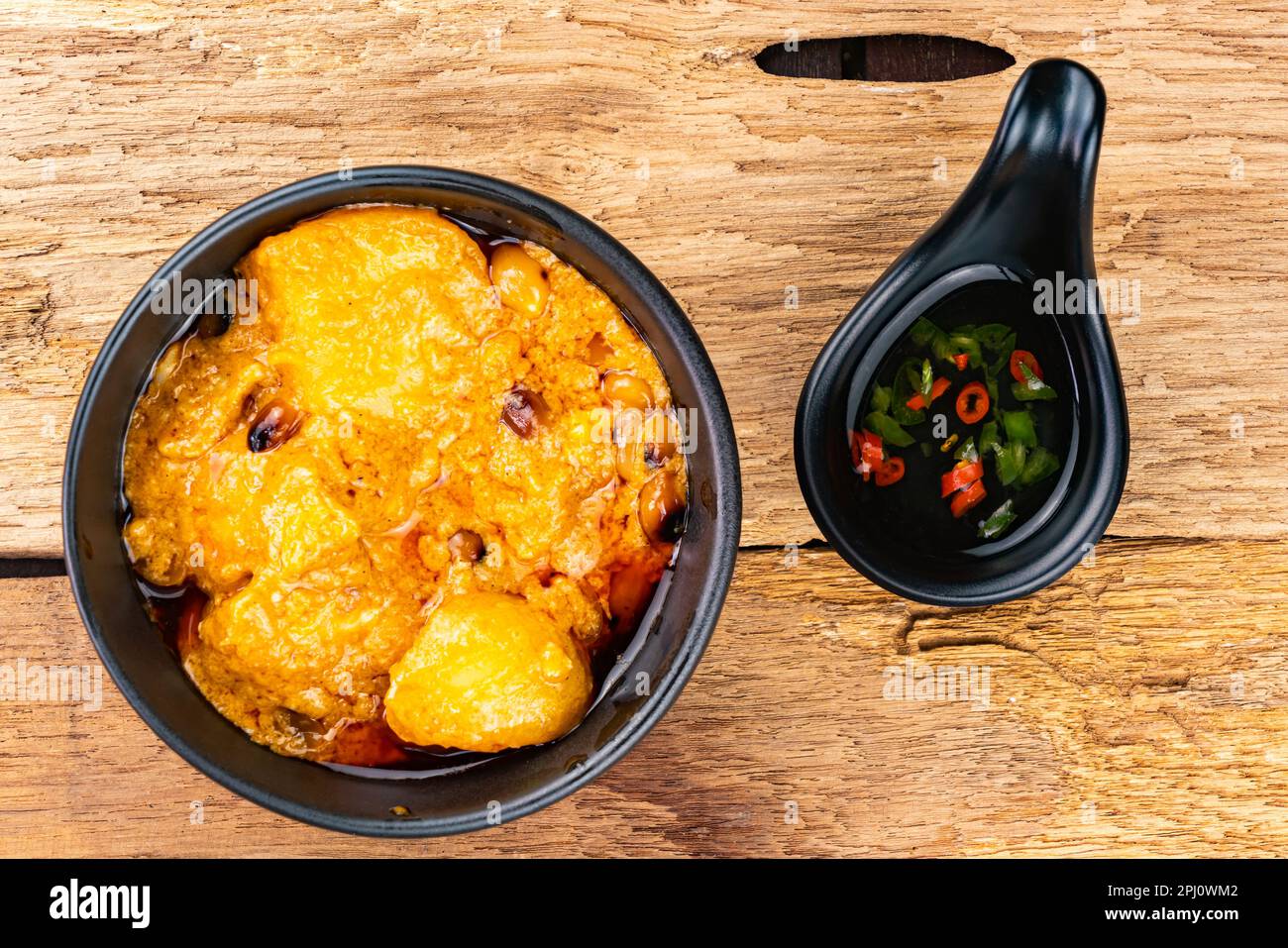 Vista dall'alto del delizioso curry di pollo speziato fatto in casa con patate, latte di cocco e peperoncino in una ciotola di ceramica nera con salsa di pesce con pezzi rossi e verdi Foto Stock