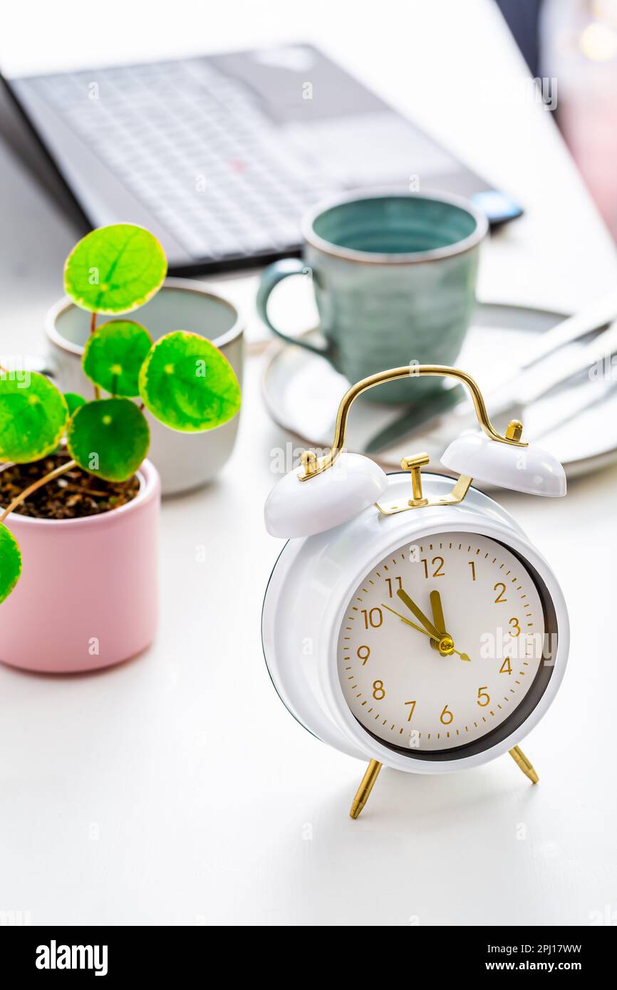 Gestione del tempo e concetto di procrastinazione. Composizione con sveglia sulla scrivania bianca, computer portatile, caffè e impianto Foto Stock