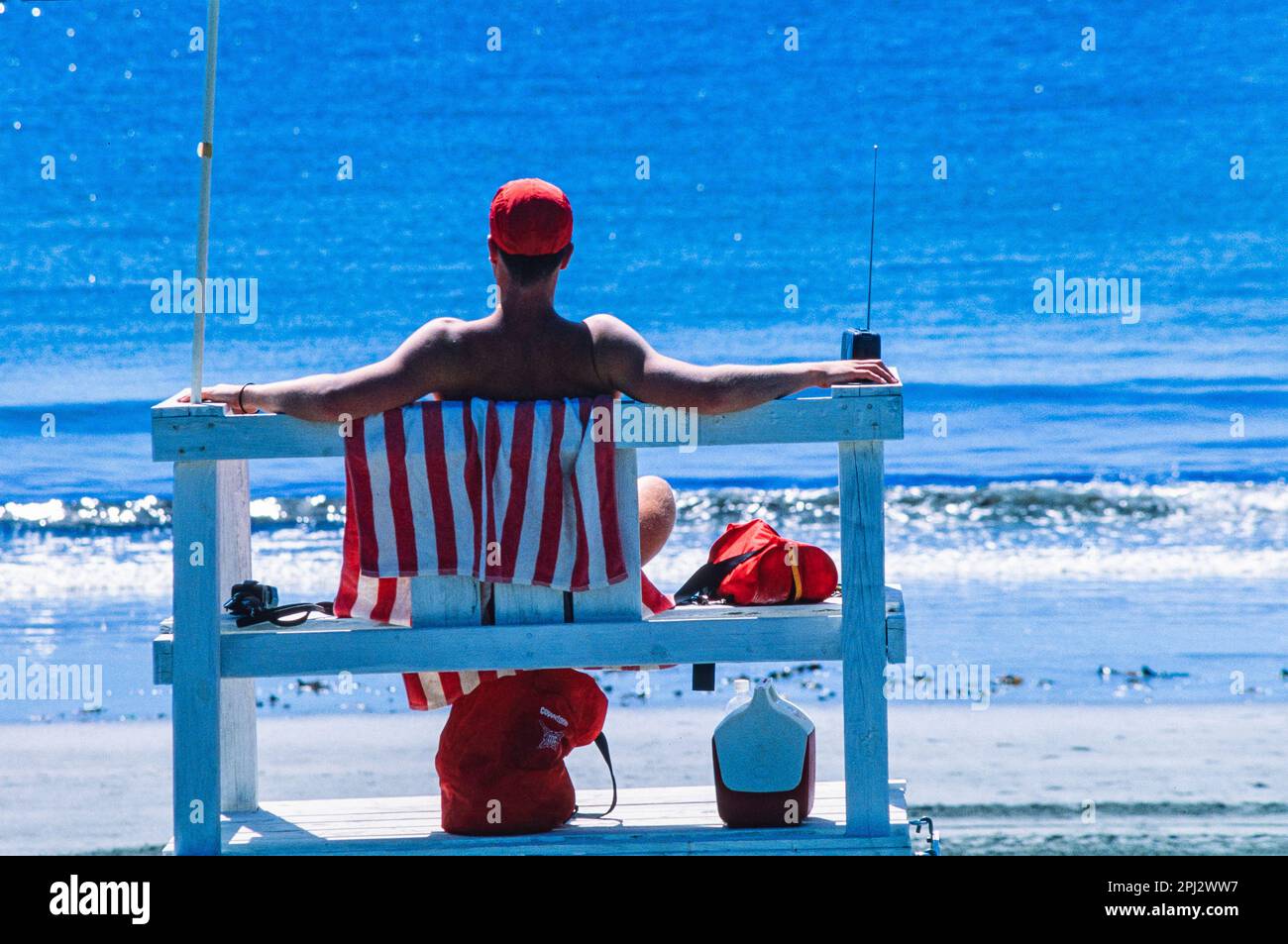 Guardia di vita con cappuccio rosso e asciugamano a strisce rosso e bianco in servizio siede sul suo persico che si affaccia su York Harbor Beach il giorno d'estate Foto Stock