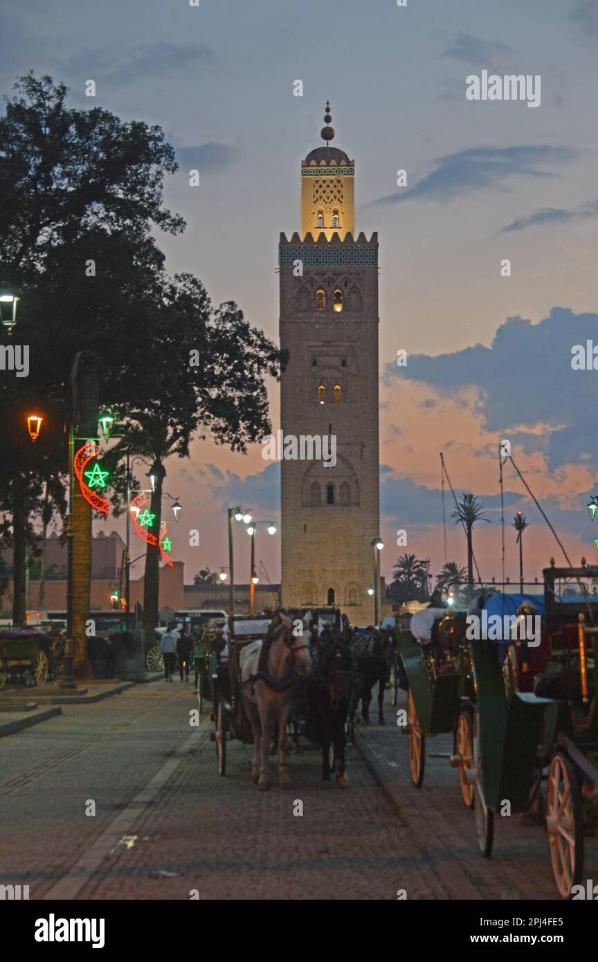 Marocco, Marakech: Il minareto illuminato della moschea di Koutoubia al tramonto, con la carrozza in piazza Jemaa el-Fnaa in primo piano. Foto Stock