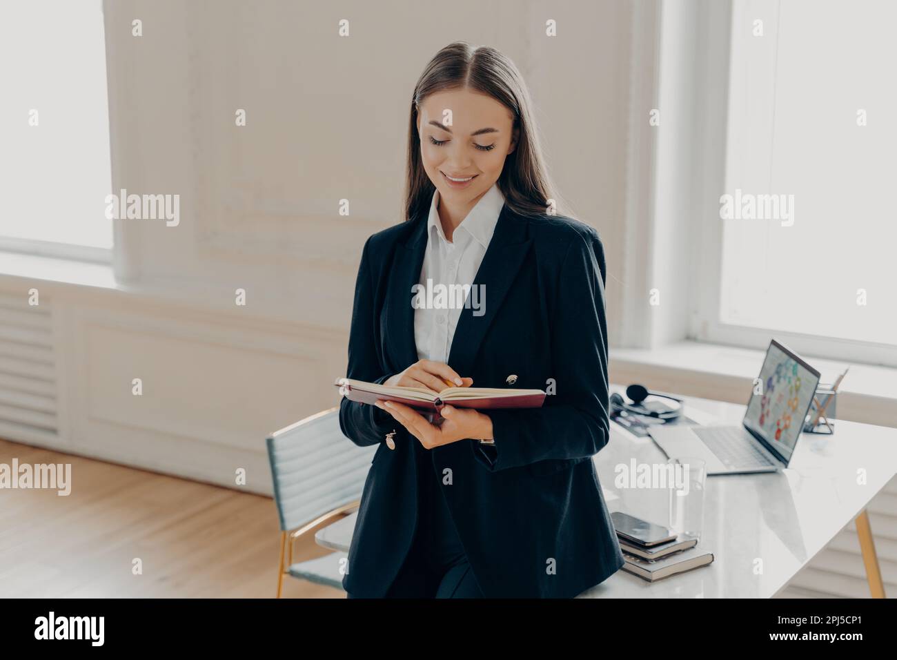 Ritratto di occupato donna d'affari con capelli dritti marrone scuro in abito nero formale scrittura e guardando il libro di note, appoggiandosi sulla scrivania laterale dentro Foto Stock