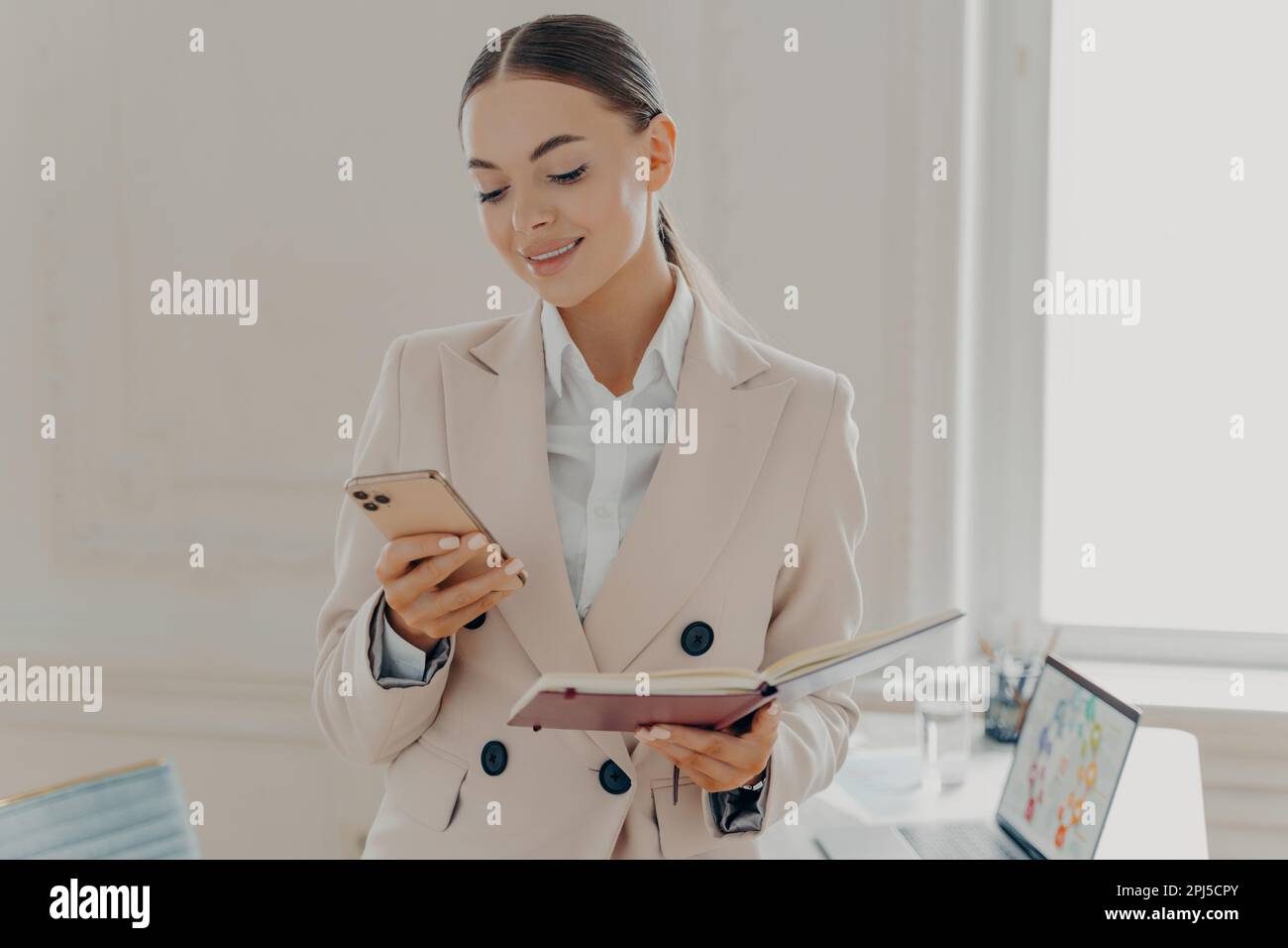 Una donna di successo che guarda lo schermo dello smartphone con il blocco note in mano, che legge le ultime notizie o controlla l'e-mail. Donna sorridente in forma beige Foto Stock