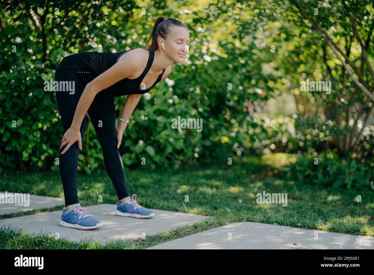 Fitness donna in nero sportsclothes mantiene le mani sulle ginocchia si appoggia in avanti esercizi all'aperto contro sfondo verde natura ascolta la musica preferita Foto Stock