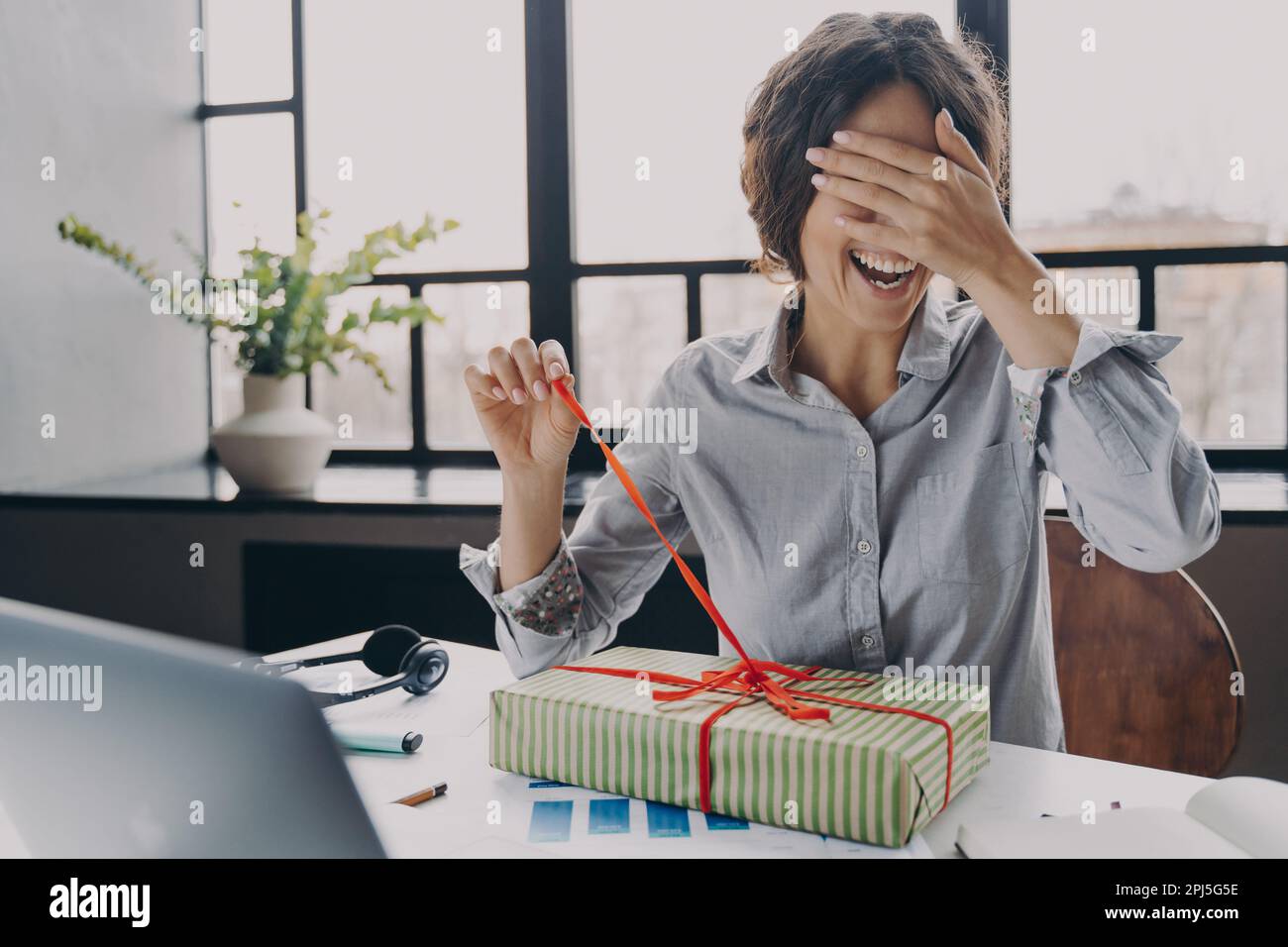 Giovane donna eccitato ufficio dipendente chiudere gli occhi con mano disimballaggio Xmas presente tirando nastro quando si siede sul posto di lavoro, felice freelance signora aperto Foto Stock