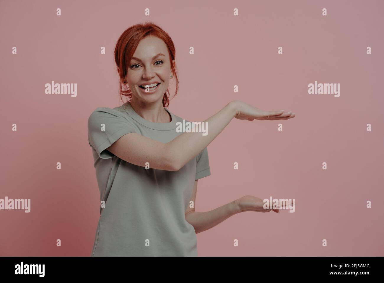 Giovane donna dai capelli rossi piacevole in t-shirt che mostra il formato enorme del prodotto o qualcosa di grande con le mani e sorridente mentre si trova isolato su backgroun rosa Foto Stock