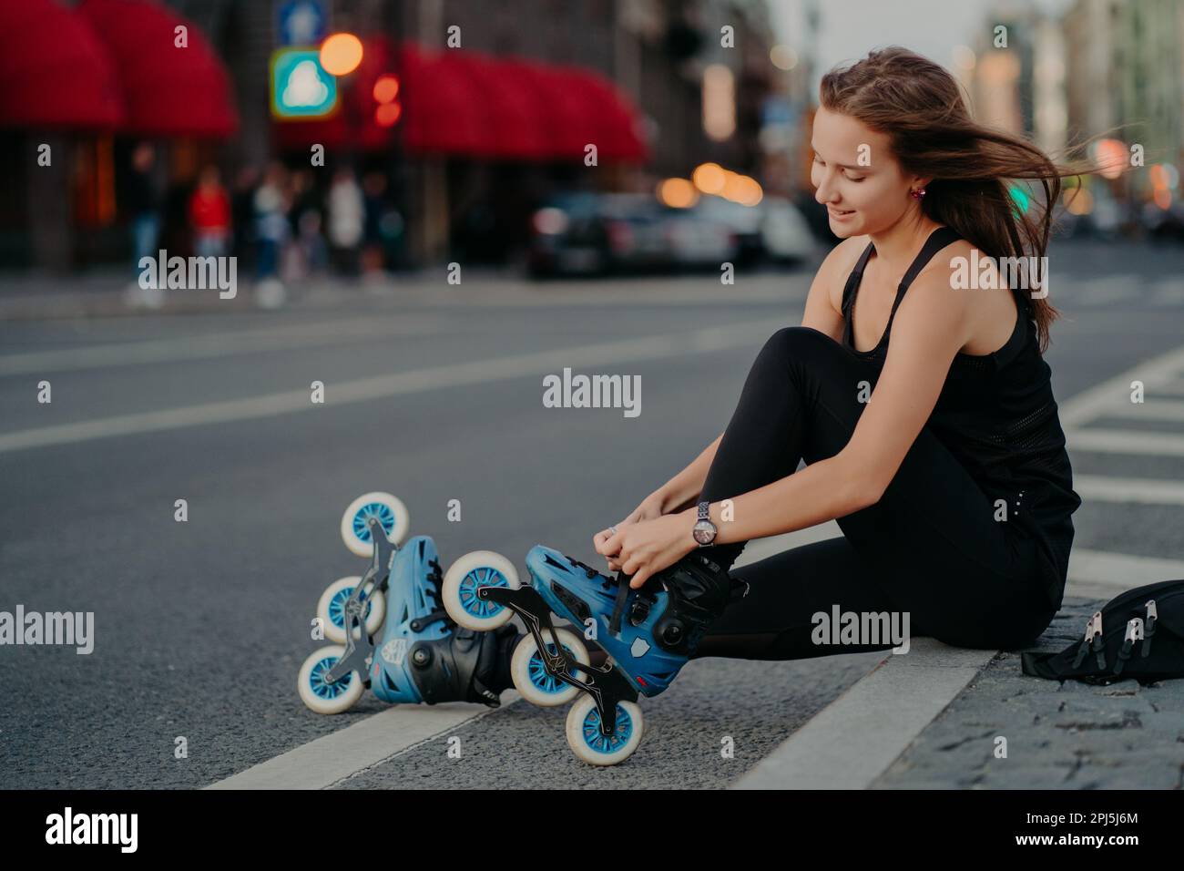 Attività all'aperto in forma stile di vita. Sideways shot di donna attiva si siede su strada regoli rollerblade si prepara per il pattinaggio mette in linea pattini ha capelli Foto Stock