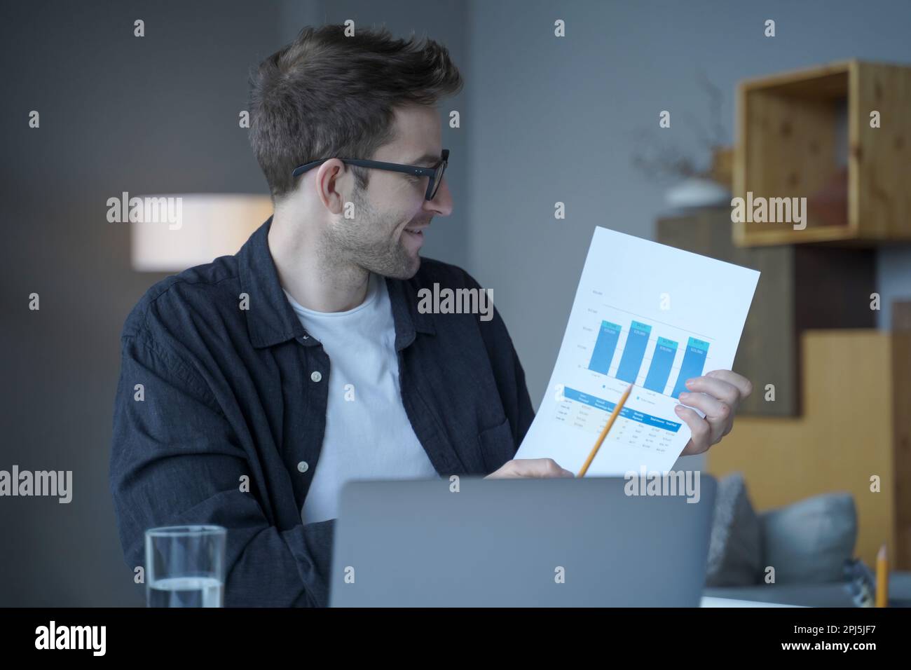 Bello imprenditore tedesco maschio soddisfatto in occhiali che rappresentano risultati e grafici di reddito sulla fotocamera laptop che punta con matita durante onlin mensile Foto Stock