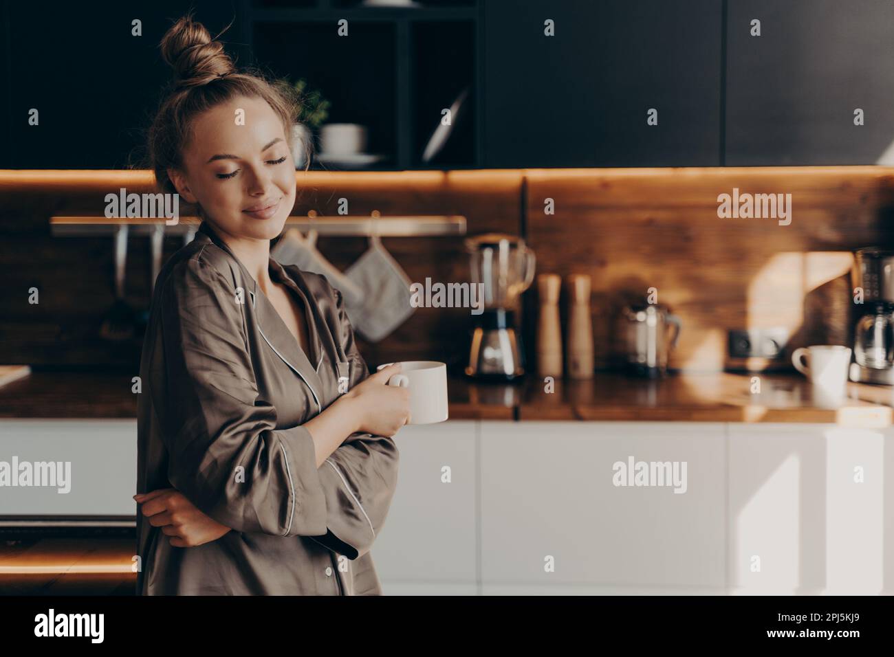 Bella giovane donna felice in piedi in pajama di seta accogliente in cucina con una tazza di caffè godendo la mattina presto tranquilla con gli occhi chiusi. Tempo libero Foto Stock