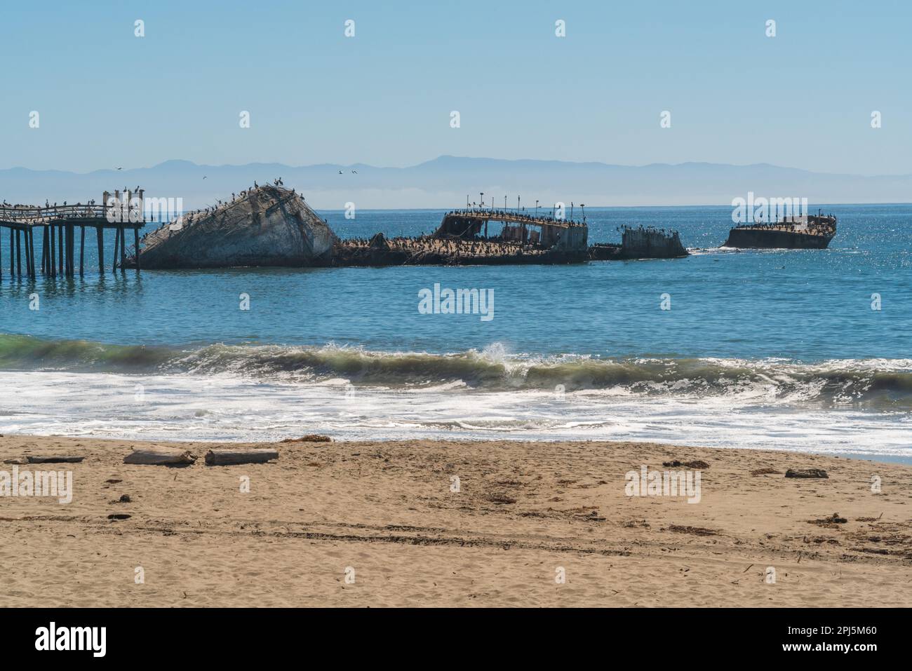 Le onde agitate si infrangono sulla spiaggia davanti al relitto e all'estremità del molo, con un debole profilo montuoso oltre, a Seacliff State Beach, California. Foto Stock