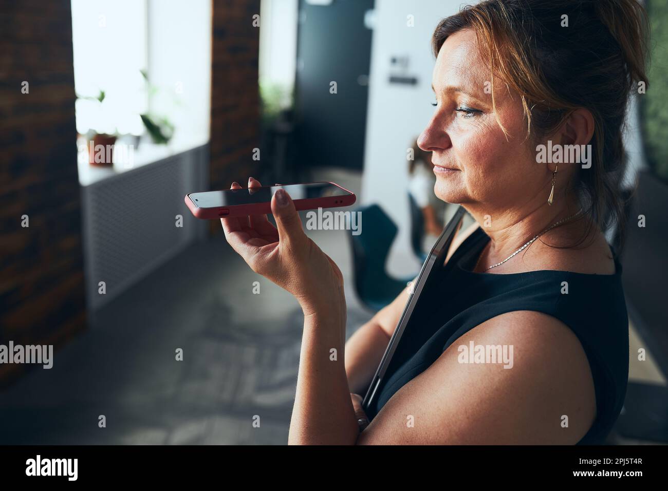 Donna che ha una chiamata di lavoro che lavora in ufficio. Donna matura che parla sul telefono cellulare utilizzando la modalità altoparlante. Utilizzando la tecnologia. Comunicazione remota. Remoto Foto Stock