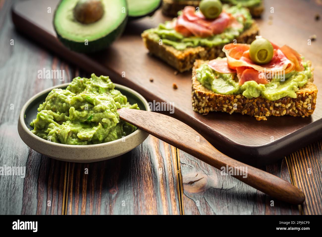 Guacamole tuffarsi o spalmare con sandwich e prosciutto aperti su tavolo da cucina in legno Foto Stock