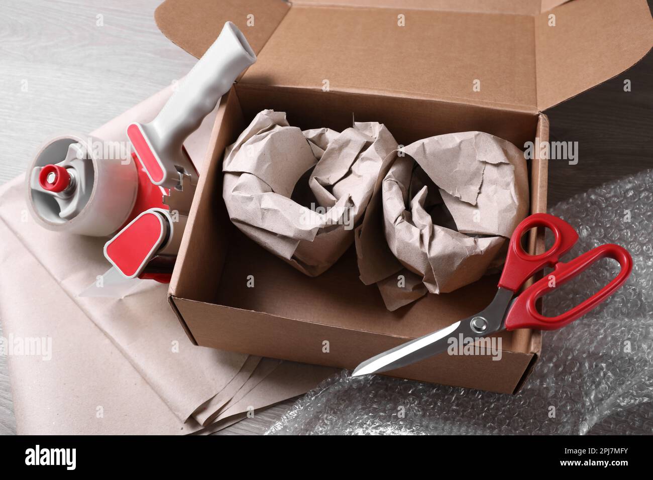 Scatola aperta con articoli avvolti, nastro adesivo, forbici, carta e pellicola a bolle su tavolo di legno Foto Stock