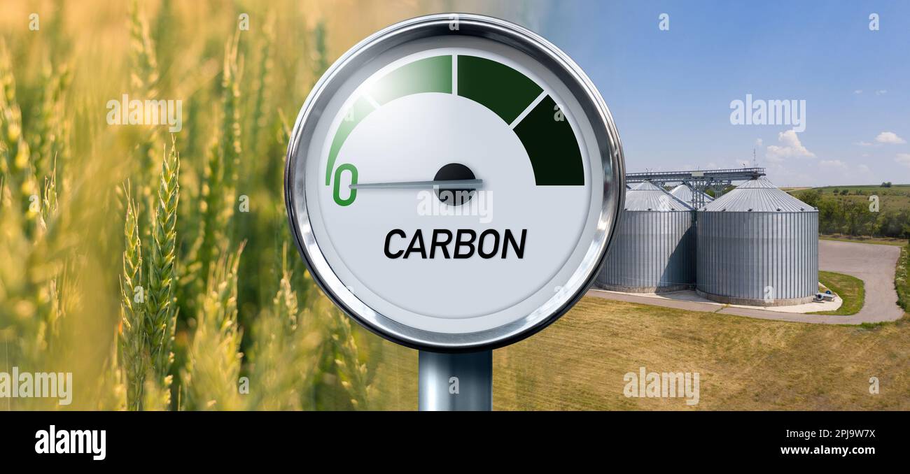 Calibro con iscrizione IN CARBONIO. La freccia punta a zero. Campo agricolo e silos su un background. Concetto di carburante biologico al carbonio neutro Foto Stock