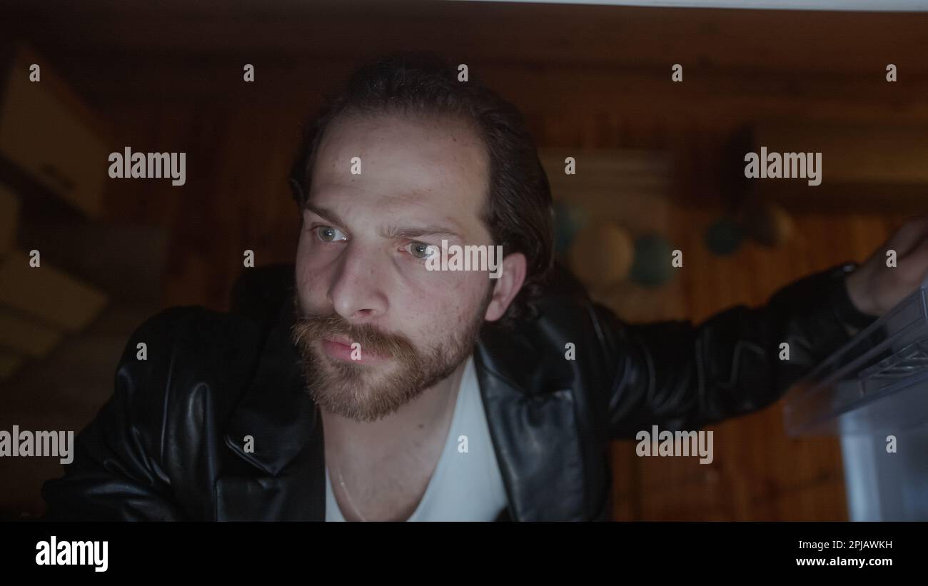 Uomo con capelli lunghi e giacca nera apre la porta del frigorifero per afferrare uno spuntino Foto Stock