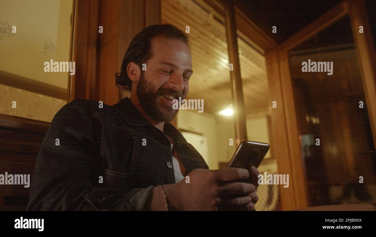 Uomo dai capelli lunghi che laughing e texting sul suo telefono dal balcone di una casa di legno illuminata da luce gialla calda Foto Stock