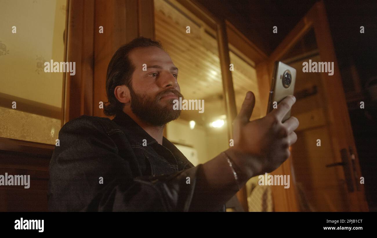 Un giovane, Bearded, Long-haired Man Video chattando sul suo telefono dal balcone di una bella casa di legno illuminata da luce gialla calda Foto Stock