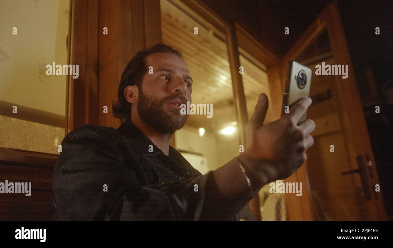 Uomo Video chattando sul suo telefono dal balcone di una bella casa di legno illuminata da luce gialla calda Foto Stock