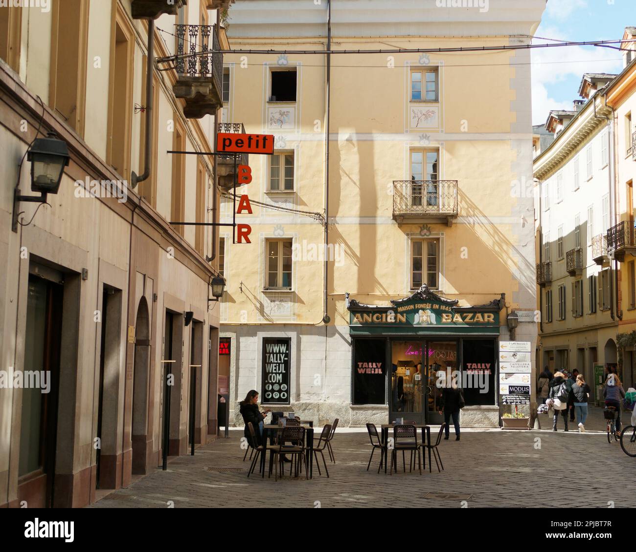 Una donna si siede fuori da un bar aka cafe in una pittoresca piazza con colori pastello e un cartello arancione, città di Aosta, Valle d'Aosta, Italia Foto Stock