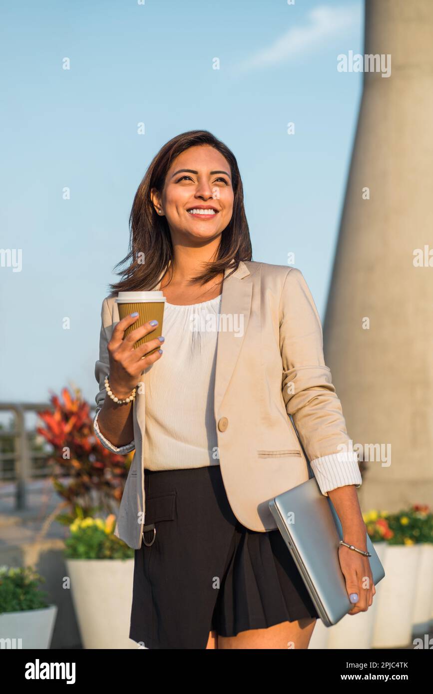 Immagine verticale di una donna d'affari Latina che guarda felicemente al futuro con un caffè e il suo computer in mano. Foto Stock