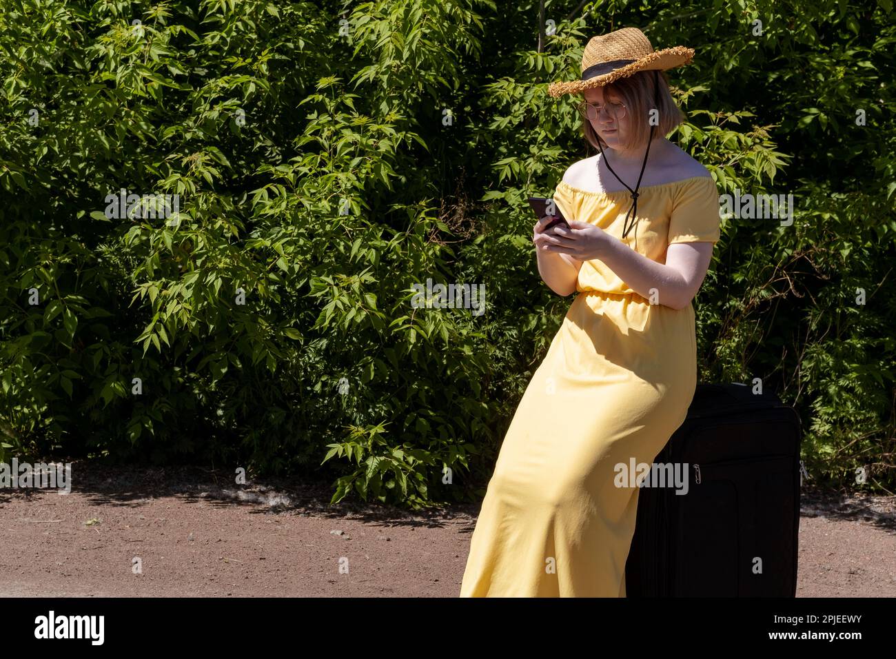 Ragazza asiatica adolescente in un vestito giallo, occhiali e un cappello seduto su una valigia e alla ricerca di un navigatore sul suo telefono. La ragazza si è persa. Ecoturismo. Foto Stock