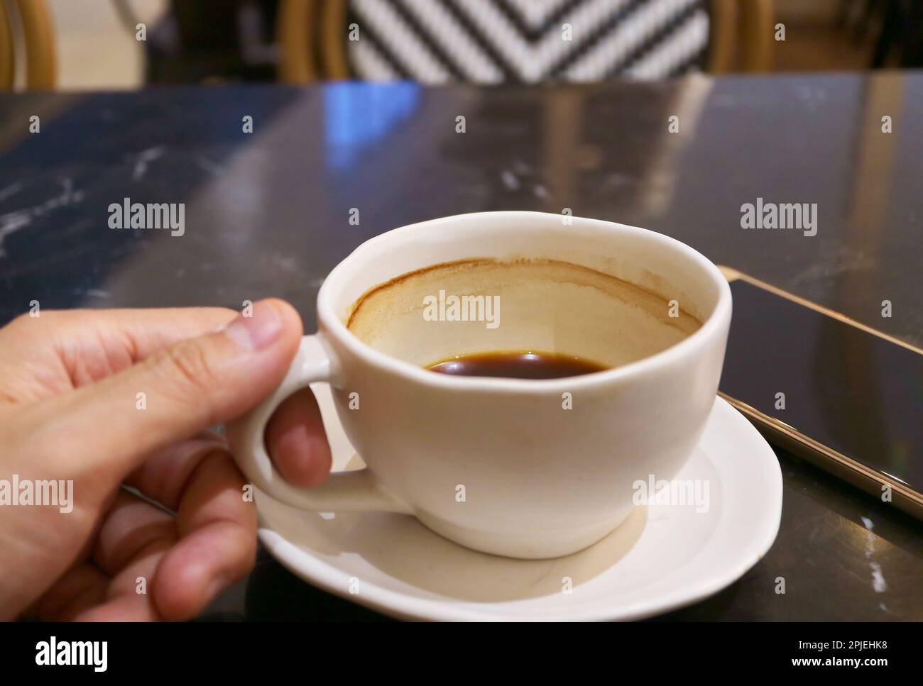 Mano che tiene una mezza tazza di caffè caldo accanto con il cellulare sul tavolo nero Foto Stock
