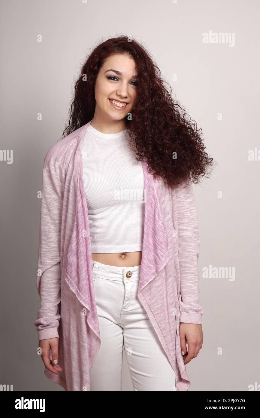 ragazza felice adolescente con lunghi capelli ricci indossando jeans bianchi e top con cardigan rosa Foto Stock
