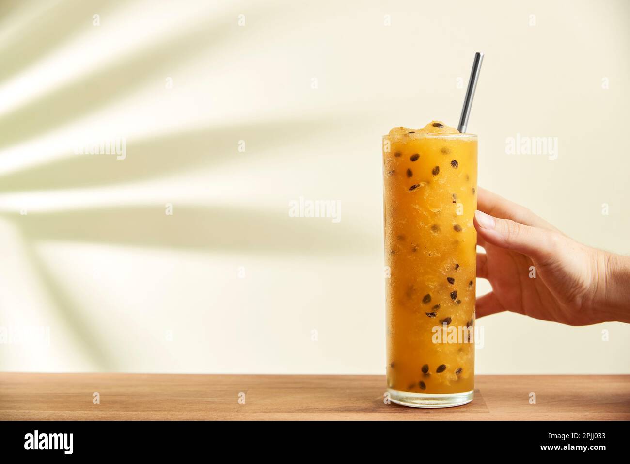 Afferrare a mano un bicchiere di una rinfrescante bevanda a base di frutto della passione, succo o cocktail, con una cannuccia ecologica riutilizzabile su una superficie di legno. L'ombra di un tr Foto Stock