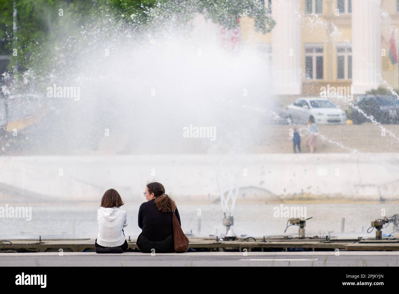 Minsk, Bielorussia - 31 maggio 2022: Le ragazze si siedono sul terrapieno sullo sfondo di una fontana funzionante che spruzzi. Schizzo della città Foto Stock