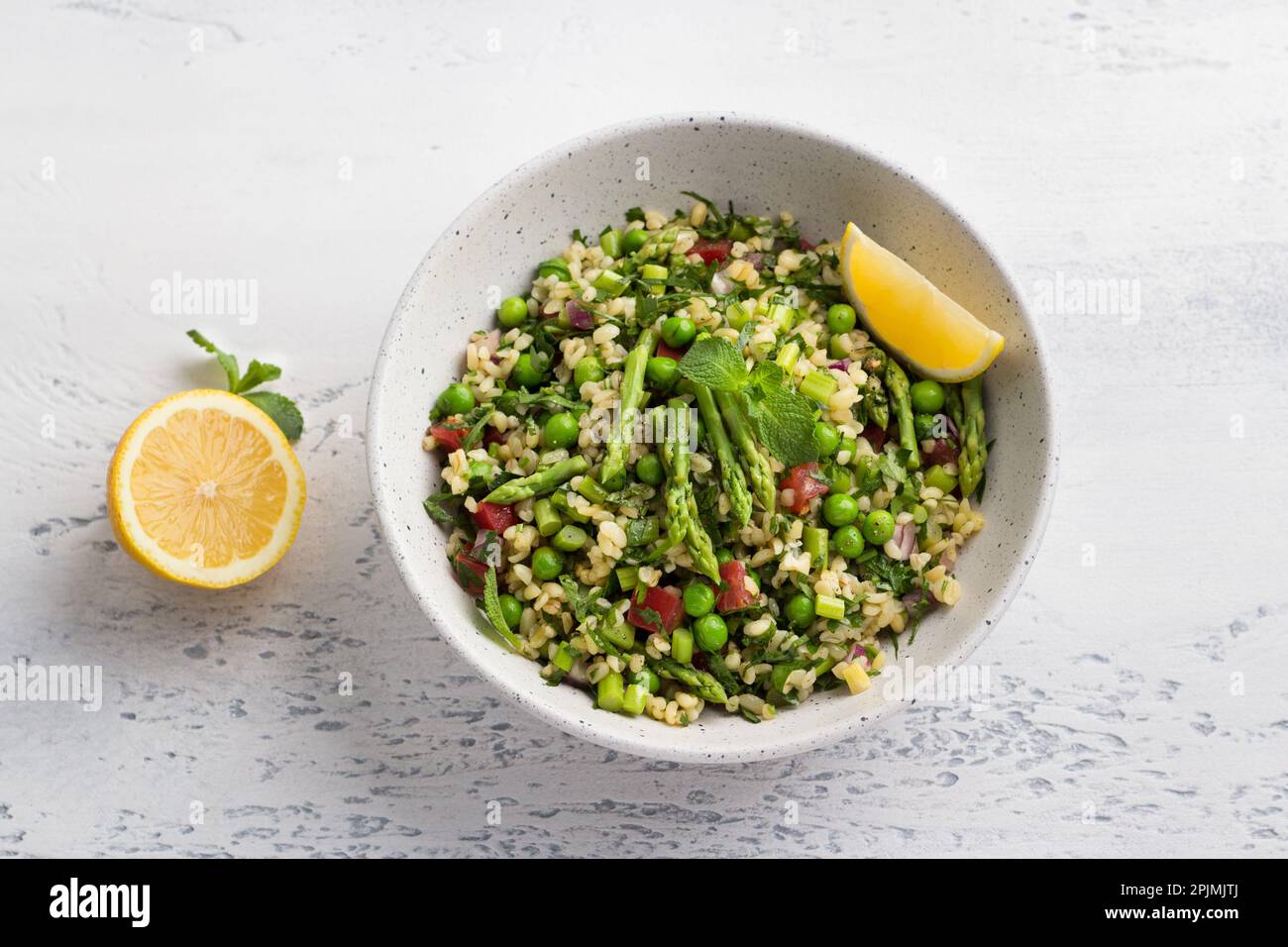 Tradizionale insalata di tableh mediorientale o araba di bulgur, pomodori, asparagi, piselli verdi e verdi con mezzo limone su un backgroun grigio chiaro Foto Stock