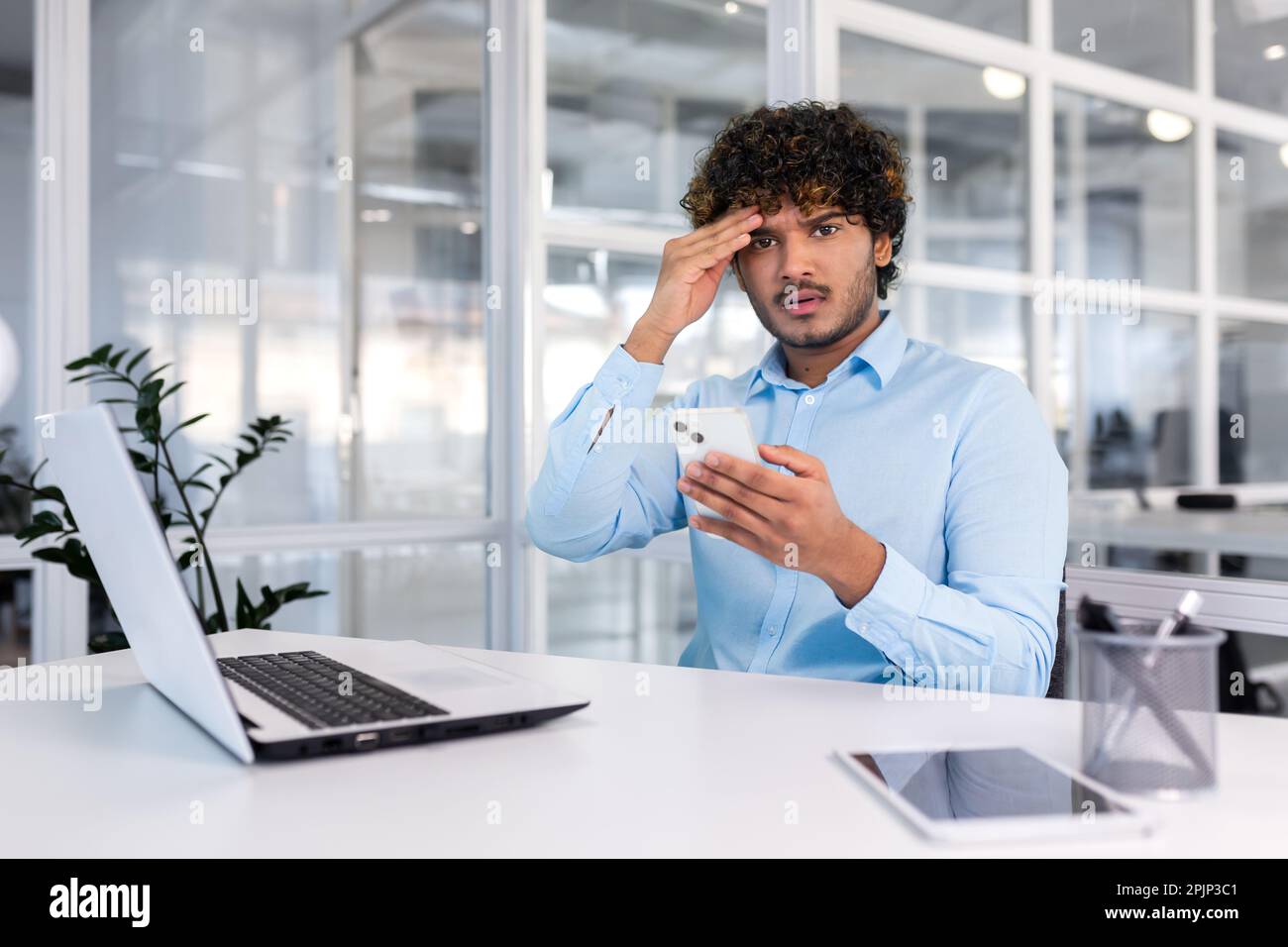 Giovane uomo d'affari ispanico seduto alla scrivania in ufficio, tenendo il telefono, tenendo la testa e guardando la macchina fotografica. ricevuto cattive notizie, messaggi, minacce, fallimento. Foto Stock