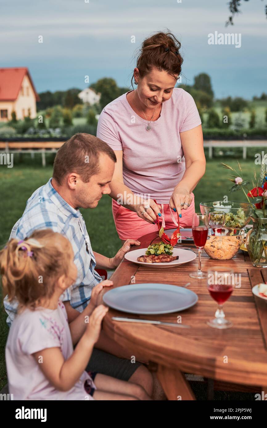Famiglia che ha un pasto dalla griglia durante la cena estiva picnic all'aperto in un giardino di casa. Primo piano di persone sedute a un tavolo con cibo e piatti Foto Stock