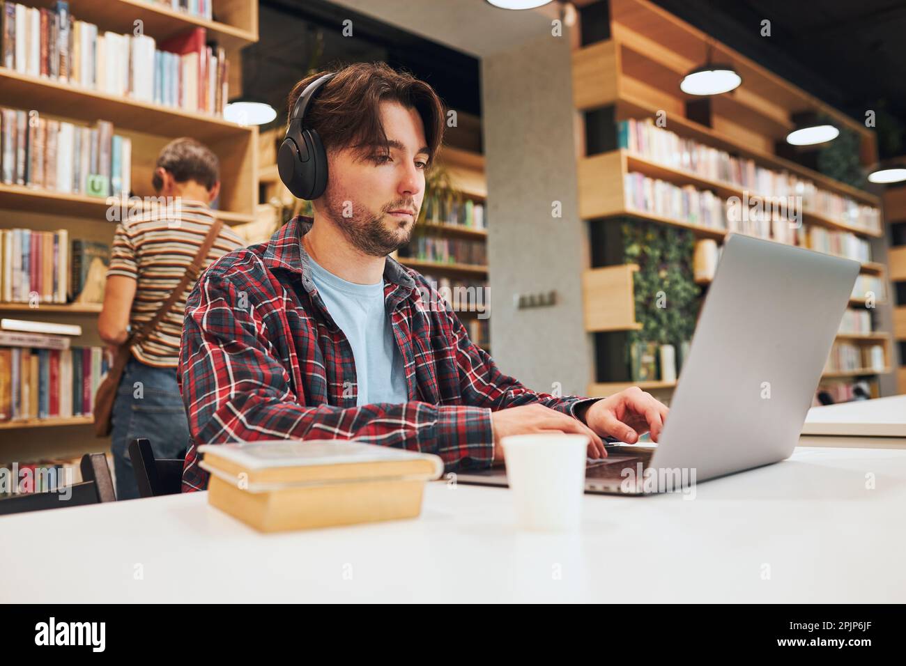 Apprendimento degli studenti nella biblioteca universitaria. Giovane uomo che si prepara per il test sul notebook. Uomo che ascolta il corso online. Studente focalizzato che studia per l'università e Foto Stock